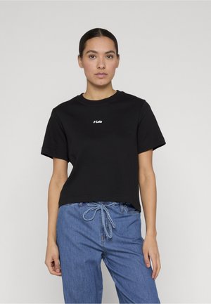 Kvinne med mørkt hår samlet bak, iført svart kortermet skjorte med teksten "Loís" og blå denim bukser med snor i midjen.