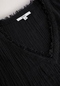 Črno oblačilo z V-izrezom in robovi z raztrganimi šivi, izdelano iz teksturiranega blaga z navpičnimi naborki. Na vrhu vidna etiketa znamke.