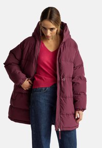 Donna che indossa un giubbotto imbottito bordeaux sopra un maglione rosso a V e jeans blu, mentre sistema la cerniera del giubbotto con una mano.