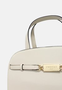 Borsa in ecopelle crema con due manici neri, dotata di una placca con logo in tonalità oro e una forma rettangolare elegante.
