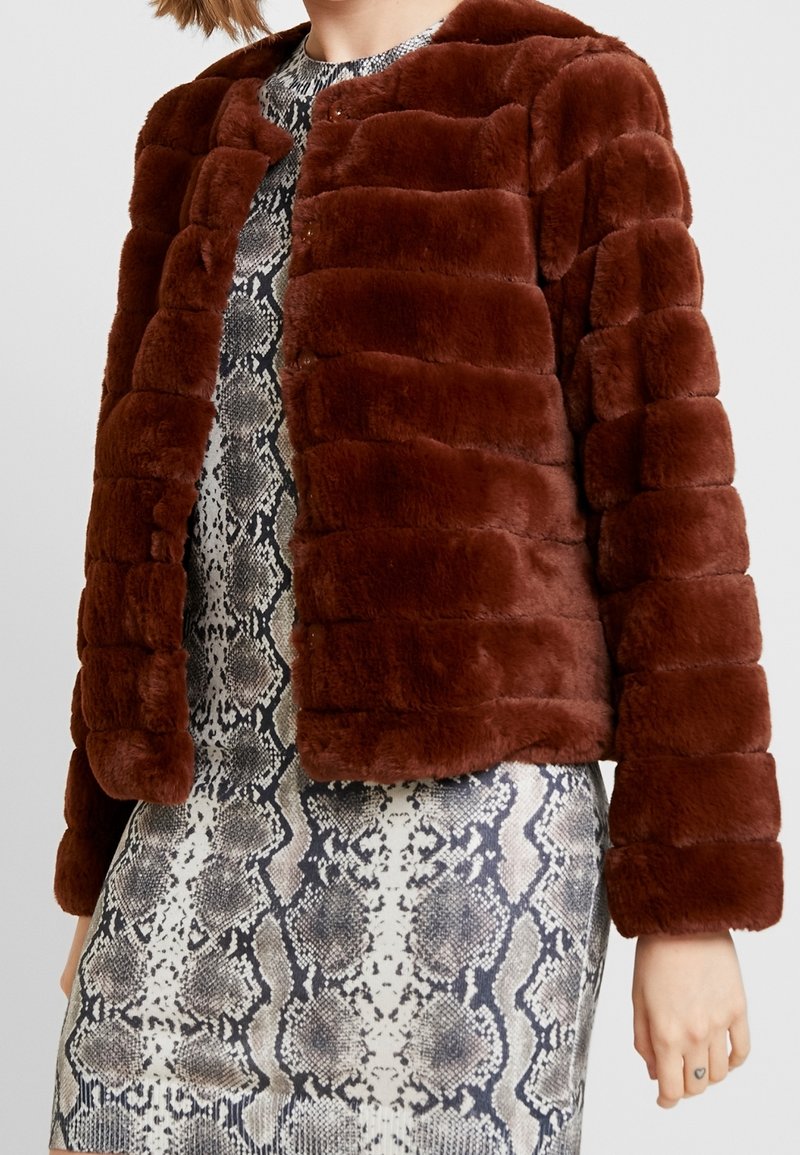 Femme portant une veste courte en fausse fourrure marron pelucheuse sur une robe noire et blanche à motif serpent, montrant un petit tatouage en forme de cœur sur son doigt.