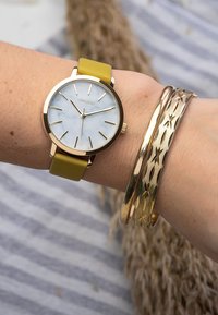 Gold watch with a white marble dial and yellow strap; accompanied by three gold bangles with varying textures and patterns on a wrist.