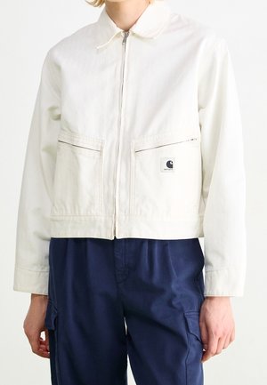 Persona con chaqueta blanca con cremallera y cuello, bolsillos delanteros y parche con logo, combinada con pantalones cargo azul oscuro.