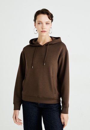 Young person with short hair wearing large hoop earrings, a brown hoodie, and dark jeans, standing against a plain white background.