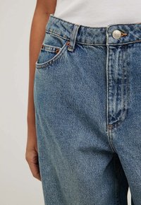 Šviesiai mėlynos džinsinės kelnės su aukštu juosmeniu, mygtukų uždarymu ir priekinėmis kišenėmis, turinčiomis išskirtinius siuvimo detalės. Minkšta, tekstūruota medžiaga.