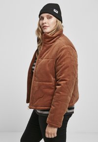 Bruine corduroyjas met een hoge kraag, gequilt design, ritsluiting en zijzakken, gedragen over een gestreept shirt. Zwarte beanie op het hoofd.