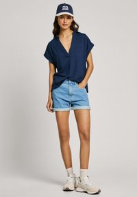 Marinblå kortärmad skjorta med krage, ljusblå denimshorts med uppvikt fåll, vita sneakers och en mörk keps med "PEPE JEANS"-logotyp.