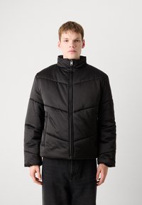 Black puffer jacket with a high collar, featuring diagonal stitching and a front zipper, shown with a model against a light background.