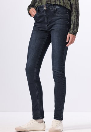 Jean skinny bleu foncé avec une texture lisse, dotés d'une fermeture à bouton à l'avant, de cinq poches et d'un design slim. Portés avec des baskets blanches.