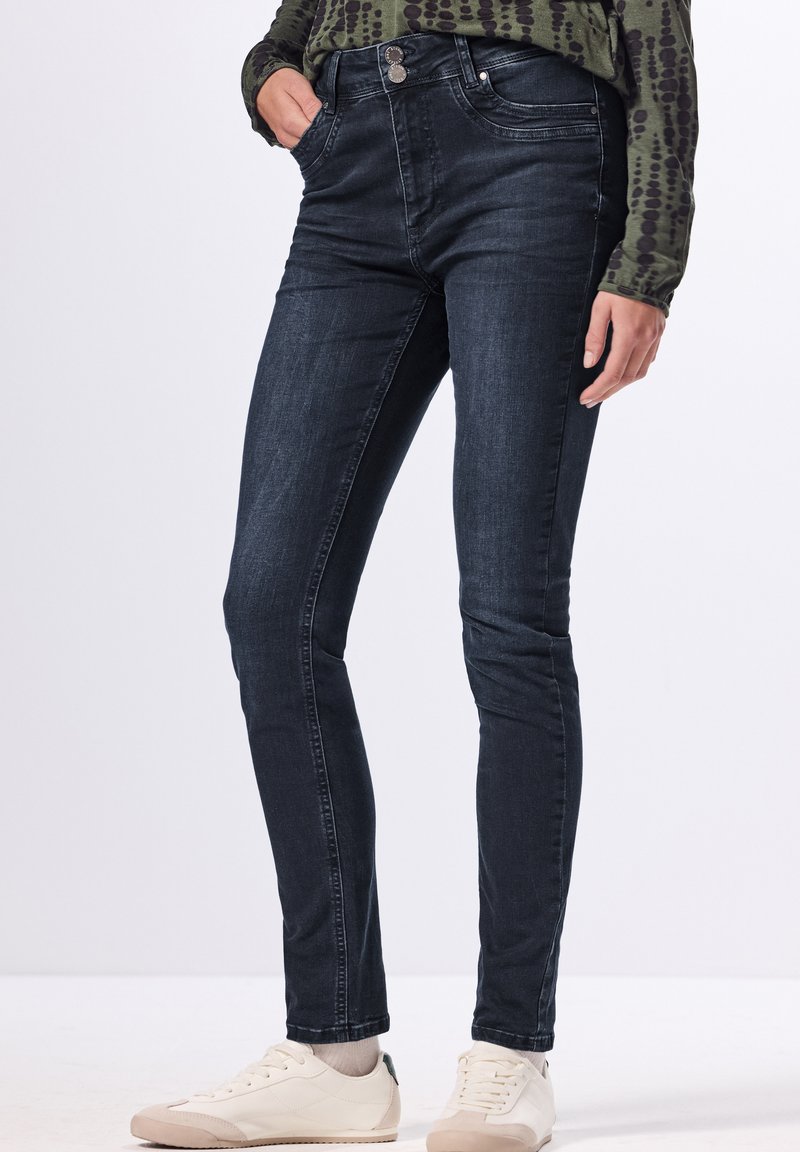 Vaqueros skinny de lavado oscuro con una textura suave, que presentan un cierre de botón en la parte delantera, cinco bolsillos y un diseño ajustado. Llevados con zapatillas blancas.