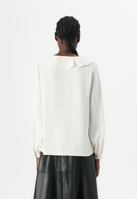 Personne aux cheveux tressés portant un chemisier blanc à manches longues avec un col à volants, associé à une jupe en cuir noire, vue de dos sur un fond blanc.