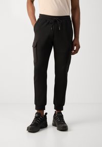 Svarte joggebukser med elastisk midje, snøring, sidelommer og avsmalnende mansjetter. Laget av mykt stoff med en jevn tekstur.