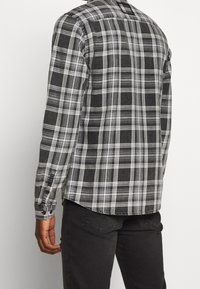 Camicia a quadri neri e grigi, maniche lunghe, con polsini a bottoni e colletto a punta. Tessuto strutturato con vestibilità rilassata e orlo curvo.