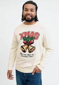Crème trui met "XMAS" in het rood, voorzien van een ontwerp van een bel en hulst. De tekst eronder luidt: "Stuur me een berichtje wanneer het Kersttijd is." Gecombineerd met een blauwe spijkerbroek.