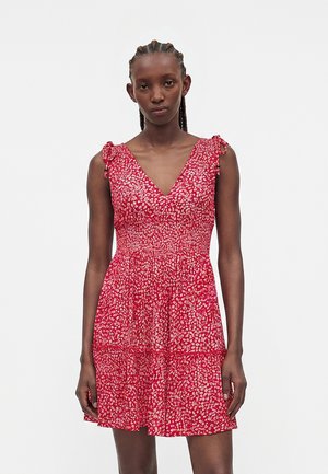 Γυναίκα με πλεγμένα μαλλιά που φοράει αμάνικο κόκκινο φόρεμα με λευκό floral μοτίβο και δεμένους ιμάντες στους ώμους, όρθια μπροστά σε απλό φόντο.