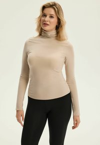 Beige Langarm-Turtleneck-Oberteil aus einem glatten, dehnbaren Stoff, mit hohem Kragen und taillierter Silhouette, kombiniert mit schwarzen Leggings.