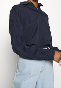 Námořnická modrá košile s dlouhým rukávem a límečkem, s uvolněným střihem a hladkou texturou, spárovaná se světle modrými kalhotami s vysokým pasem.