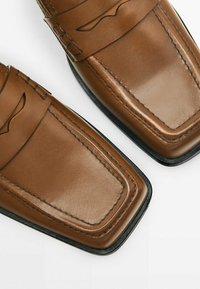 Mocassins en cuir marron avec des bouts carrés et des semelles noires, dotés d'une sangle décorative et de détails de couture.