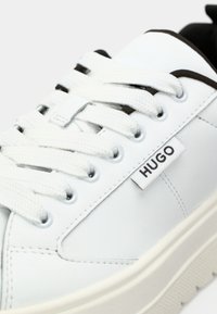 Białe skórzane trampki o strukturalnej powierzchni, okrągłym nosku i grubej białej podeszwie. Posiadają płaskie białe sznurówki oraz czarny akcent na języku. Marka: HUGO.