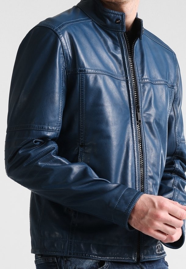 Homme portant une veste en cuir bleu foncé zippée avec un col à boutons-pression, mains jointes, sur un fond clair uni.