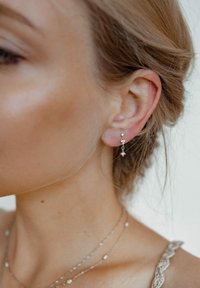 Srebrne kolczyki z trzema małymi, przezroczystymi kamieniami szlachetnymi, o wiszącym designie. Kolor skóry jest jasny. Włosy ułożone w luźne fale.