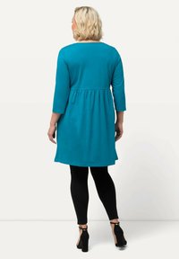 Tyrkysová tunika s třičtvrtinovými rukávy, elastickým pasem a rozšířeným spodním lemem, spárovaná s černými legínami a sandály na podpatku.