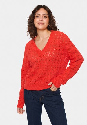 Red knitted sweater with a V-neck, featuring an openwork pattern and ribbed cuffs. Paired with dark denim jeans, showcasing a casual style.