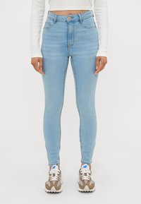 Lyseblå høytlivs skinny jeans laget av denim, med fem lommer og knappelukking. Modellen har på seg nøytrale sneakers.