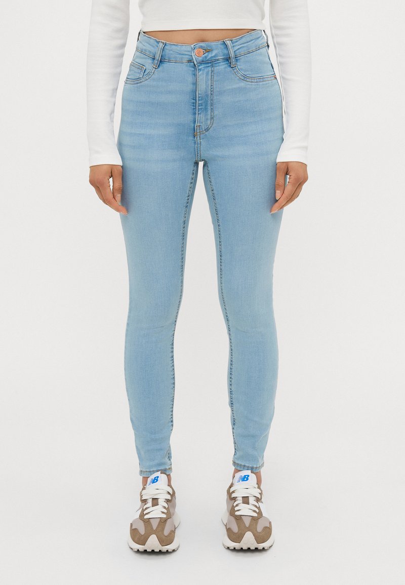 Lyseblå høytlivs skinny jeans laget av denim, med fem lommer og knappelukking. Modellen har på seg nøytrale sneakers.