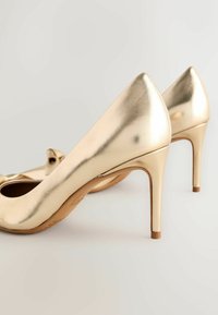 Zelta augstpapēžu kurpes ar gludu, spīdīgu apdari, ar smailu purngalu un smalku loku detaļu priekšpusē, uz šauriem stiletto papēžiem.