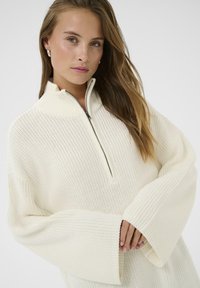 Pull en maille crème avec demi-zip, manches larges et texture côtelée. Dispose d'un col haut et d'une coupe décontractée.