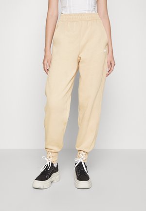 Beige sweatpants with elastic waistband, cuffed ankles, soft texture, and white logo detail. Paired with black sneakers and white laces.