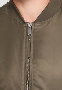 Olijfgroene bomberjas met een gladde, glanzende textuur, geribde kraag en een frontale ritssluiting met zilverkleurig beslag.