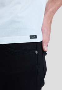 Close-up of a person wearing a white t-shirt with a small "Lee" tag and black jeans, with hand resting near the pocket.