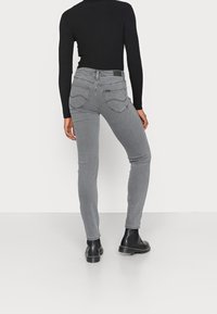 Persona que lleva vaqueros ajustados de cintura alta en gris, una camiseta de manga larga de canalé negra y botas negras hasta el tobillo, de pie contra un fondo blanco liso.