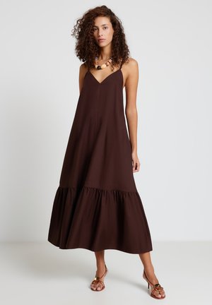 Donna con capelli ricci che indossa un abito lungo marrone scuro senza maniche e una collana con grosse perle, in piedi davanti a uno sfondo semplice.