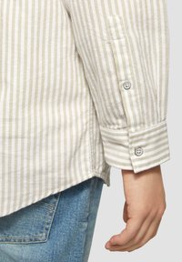 Gestreept overhemd met knopen in beige en wit, voorzien van een manchet met dubbele knoopsluiting. Het materiaal lijkt licht en heeft een gladde textuur.