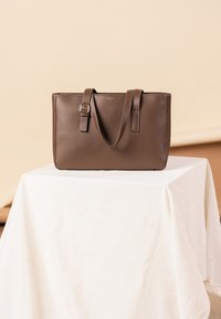 Brown leather tote bag with two sturdy handles, featuring a smooth texture and a subtle metallic buckle detail on one handle.