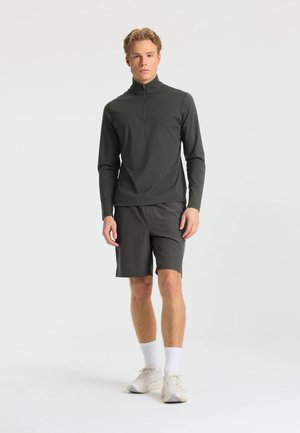 Ung mand står iført en mørk langærmet lynlås-trøje, mørke shorts, hvide sokker og lyse sneakers foran en ensfarvet baggrund.