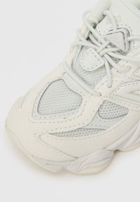 Biele a svetlošedé tenisky zo sieťoviny a kože, zaobleného tvaru, s textúrovanými akcentmi a šnúrkami. Viditeľný detail loga.