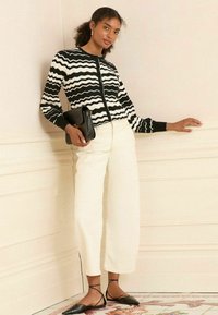 Czarny i biały kardigan w wzór zygzak, dopasowany krój, w zestawieniu z kremowymi spodniami w szerokim kroju i czarnymi czółenkami ze spiczastym noskiem. Trzyma czarną kopertówkę.