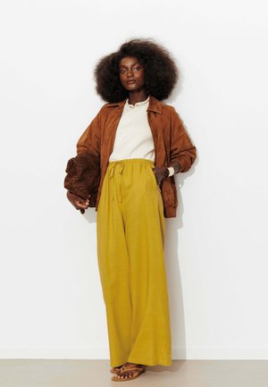 Femme aux cheveux bouclés volumineux, debout contre un mur blanc, portant une veste marron, un haut blanc, un pantalon large jaune, tenant un chapeau à imprimé léopard.