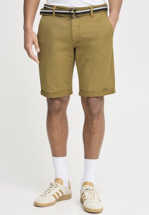 Shorts en coton kaki avec des revers, dotés d'une ceinture rayée noir et blanc. Portés avec des chaussettes blanches et des baskets beiges.