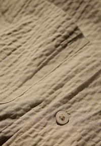 Teléfono beige arrugado con un cierre de botón y una textura distintiva. Presenta un detalle de bolsillo y líneas de costura sutiles.