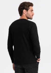 Uomo con capelli castani che indossa una maglietta nera a maniche lunghe e jeans scuri, in piedi con le mani nelle tasche, rivolto a destra su uno sfondo semplice.