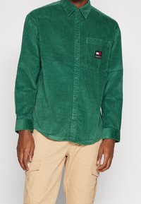 Zelená manšestrová košile s límečkem na knoflíky, žebrovanou texturou, přední kapsou s logovou nášivkou a dlouhými rukávy.