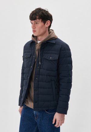 Granatowa pikowana kurtka puchowa z kołnierzem, dwoma kieszeniami na piersi i zapięciem na zatrzaski. Nosi na sobie beżowy hoodie i niebieskie dżinsy.