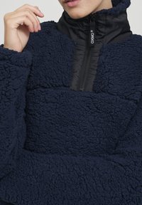 Pullover in pile blu navy con colletto in nylon nero a mezzezip. Presenta una superficie soffice e texturizzata e una toppa liscia a contrasto sul petto.