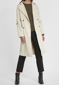 Manteau beige à double boutonnage, col en pointe et deux poches latérales, porté par-dessus un haut en maille vert olive et un pantalon noir.