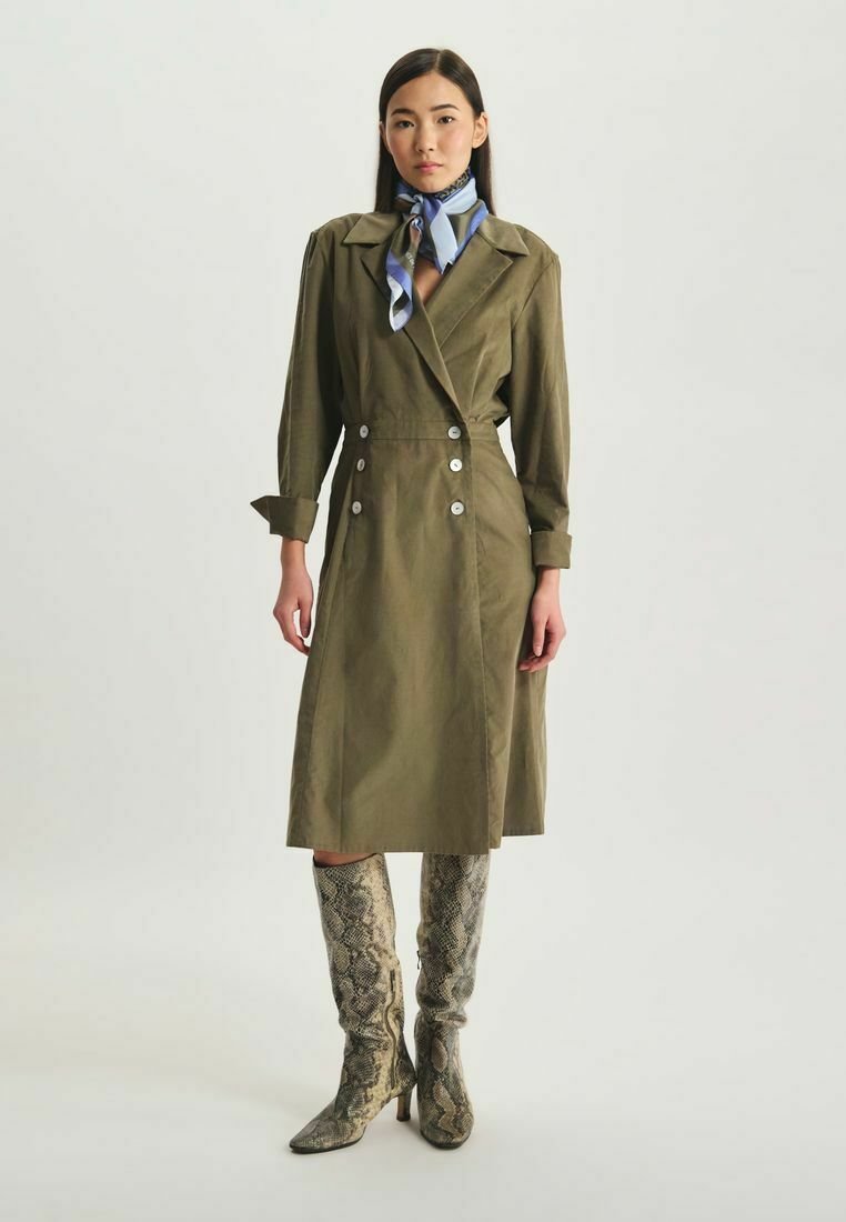 Manteau trench vert olive avec des boutons croisés, des manches longues et une ceinture à nouer, associé à des bottes jusqu'aux genoux à imprimé serpent et à une écharpe bleue.
